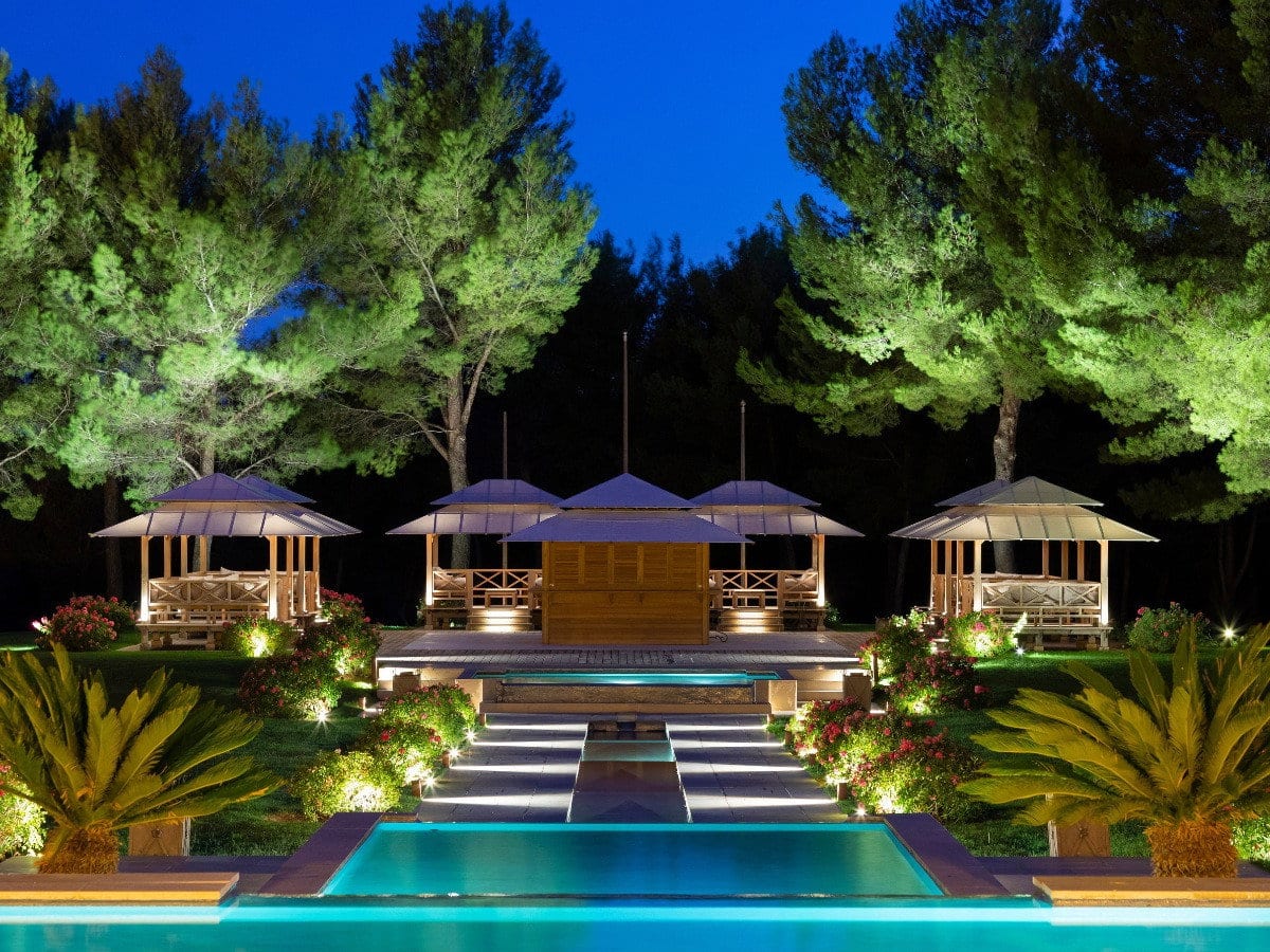 Pines and cycads uplit around a resort pool, reflected in the water