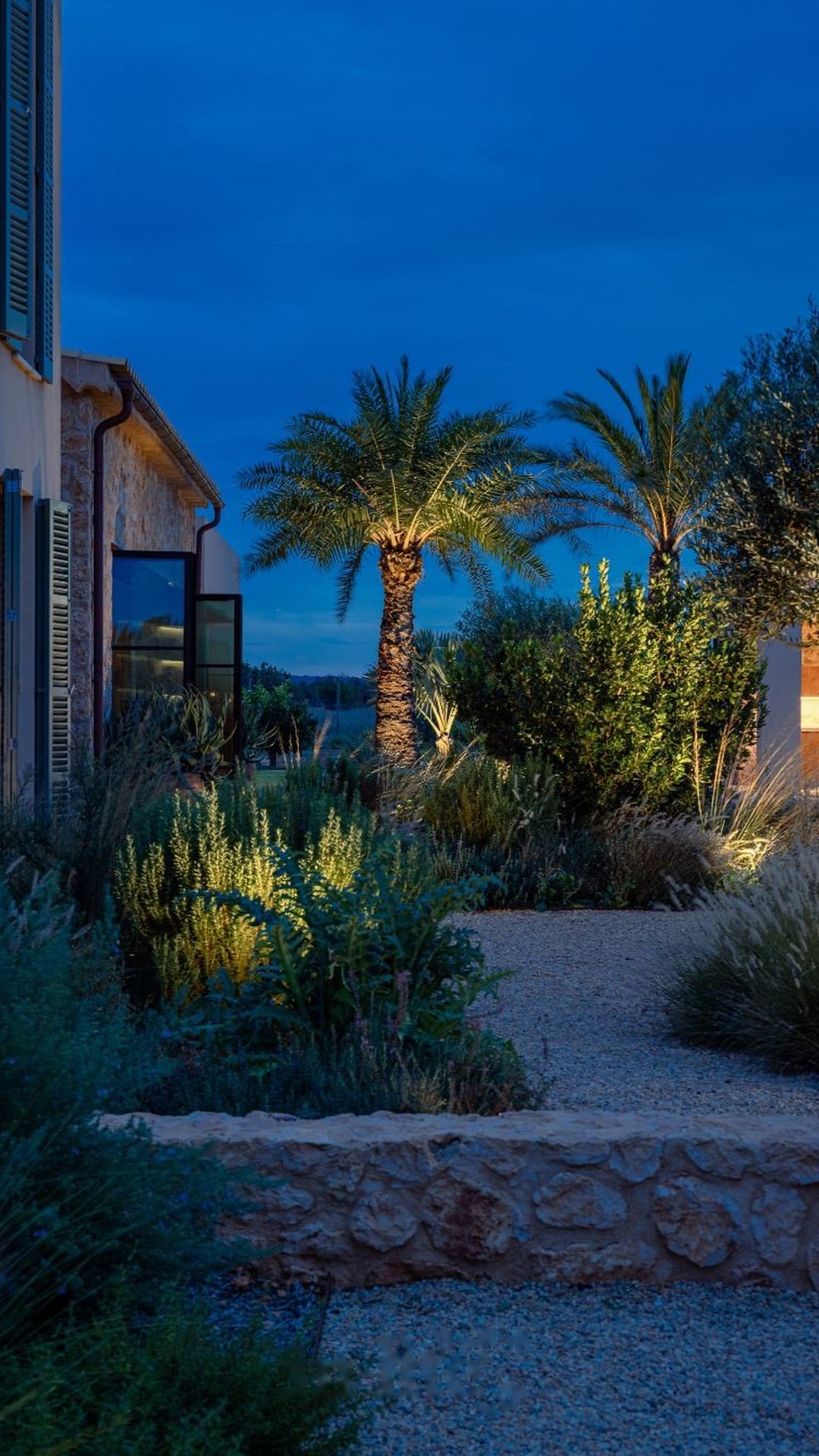 Palmeras y muro de piedra seca suavemente iluminados en un jardín costero de Mallorca